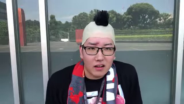 室内でモヒカン型の被り物を着け、赤と白のマフラーを掛けて話すみつお