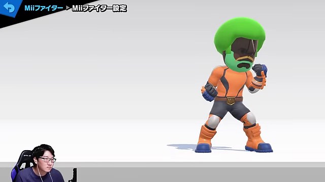 スマブラSPのMiiファイター編集画面。緑の髪とオレンジの服のMiiが立ち、みつおのカメラ映像が左下にある
