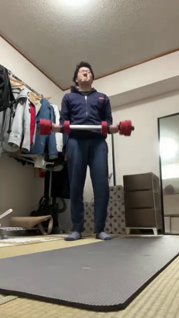 みつおがマットの前に立ち、赤い重りを付けた1本のバーを腰の高さに持ち上げている公開ライブの場面