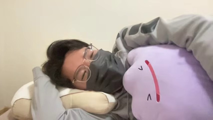 布団でクッションを抱え、頭に手を置いて大会の登録名を話すみつお
